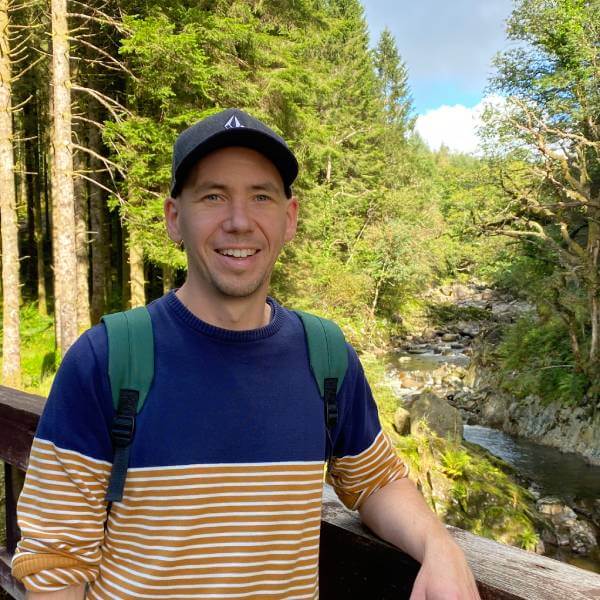 Dave is on a scenic forest trail, leaning against a wooden railing. Behind him, a rocky stream winds through lush green trees under a bright summer blue sky. He wears a navy and mustard-striped sweater and a black cap, looking relaxed and happy outdoors.